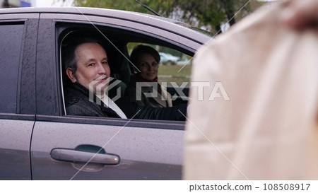 Man receives cardboard bag with meal from delivery person sitting in car with wife driver. Man content with food delivered to car. Cheerful man awaiting food delivery from courier sitting in car Man receives cardboard bag with meal from delivery person sitting in car with wife driver. Man content with food delivered to car. Cheerful man awaiting food delivery from courier sitting in car 108508917