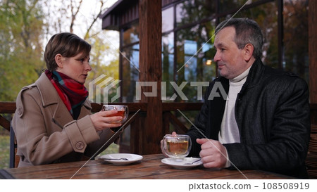 Tea drinking of man and woman talking at business meeting in park cafe. Meeting for negotiations and tea in park between man and woman. Man and woman discuss plans drinking tea at meeting in park Tea drinking of man and woman talking at business meeting in park cafe. Meeting for negotiations and tea in park between man and woman. Man and woman discuss plans drinking tea at meeting in park 108508919