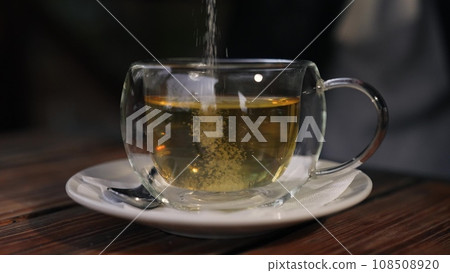 Person adding sugar into green tea in transparent cup at wooden table in housing. Moment captures human relaxation as sweetens tea in comfort of home. Human enjoys warm tea with sugar Person adding sugar into green tea in transparent cup at wooden table in housing. Moment captures human relaxation as sweetens tea in comfort of home. Human enjoys warm tea with sugar 108508920