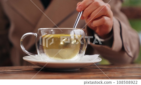 Woman stirs green tea with spoon in glass cup in cafe of park. Woman takes tea break with rotating spoon to stir sugar in cafe. Tea for maintaining woman figure with stirring spoon in cafe 108508922