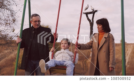 Parents swing son on swings infusing surroundings with joy in park. Parents amid kaleidoscope of colours with son share moments. Parents swinging son on swing on park playground enjoying experience 108508932