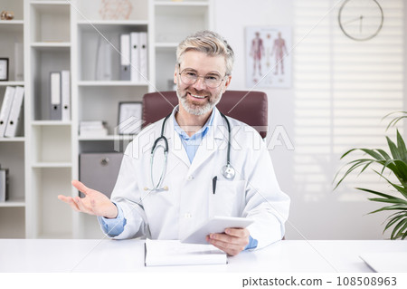 Mature experienced doctor with gray hair, smiling and looking at camera, consulting patients online remotely, senior man working remotely at workplace inside office. 108508963