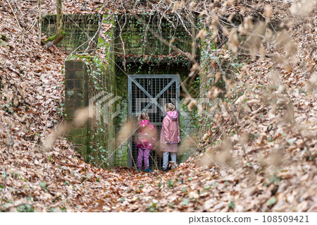 Two little girl friends kids stay at entrance of old abandoned entrance door of scary underground bunker shelter. Lost run away children in forest woods foliage danger secret building entry Two little girl friends kids stay at entrance of old abandoned entrance door of scary underground bunker shelter. Lost run away children in forest woods foliage danger secret building entry 108509421