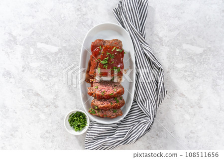 Flat lay. Slicing classic meatloaf with a sweet glaze on a white serving plate Flat lay. Slicing classic meatloaf with a sweet glaze on a white serving plate 108511656