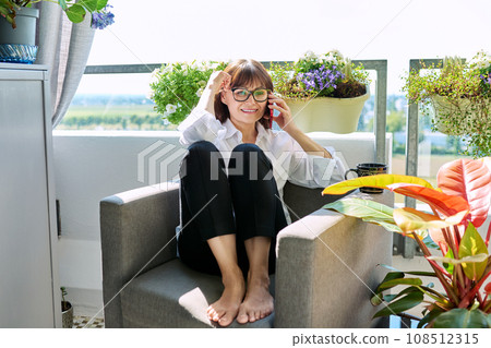 Middle aged happy woman sitting at home in chair with cup talking on mobile phone Middle aged happy woman sitting at home in chair with cup talking on mobile phone 108512315