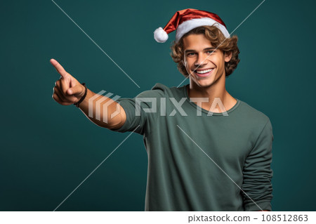 Young man in Santa hat, smiling guy pointing finger to side. Portrait of handsome person isolated on blue green studio background. Concept of Christmas, show, welcome, xmas, gif 108512863