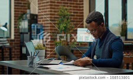 African american man checking results, reviewing data statistics on documents in coworking space. Analyzing research information to create business presentation for boardroom meeting. 108513801