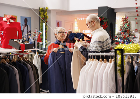 Joyful old people comparing items in store, showing each other trendy jackets to buy for festive christmas dinner. Senior clients smiling while shopping for clothes on discount, xmas decor. 108513915