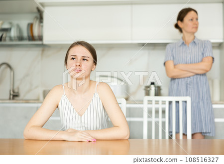 Offended young girl sitting at the kitchen table, angry mother standing behind Offended young girl sitting at the kitchen table, angry mother standing behind 108516327