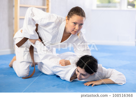 Sportive woman holds her opponent by force while sitting on mats during judo classes Sportive woman holds her opponent by force while sitting on mats during judo classes 108516368