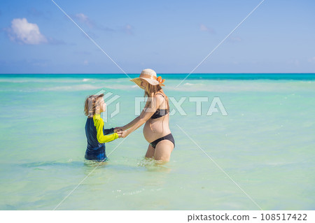 A radiant pregnant mother and her excited son share a tender moment on a serene, snow-white beach, celebrating family love amidst nature's beauty A radiant pregnant mother and her excited son share a tender moment on a serene, snow-white beach, celebrating family love amidst nature's beauty 108517422
