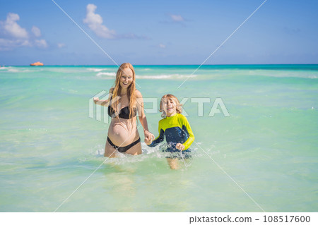 A radiant pregnant mother and her excited son share a tender moment on a serene, snow-white beach, celebrating family love amidst nature's beauty A radiant pregnant mother and her excited son share a tender moment on a serene, snow-white beach, celebrating family love amidst nature's beauty 108517600