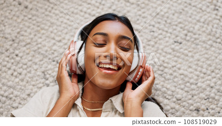Headphones, happy and young woman on the carpet on the floor listening to music or album. Smile, calm and Indian female person streaming podcast, radio or playlist and chilling in living room of home 108519029