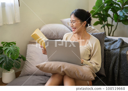 Happy Asian woman is working from home, researching information in a book, and working on her laptop Happy Asian woman is working from home, researching information in a book, and working on her laptop 108519668