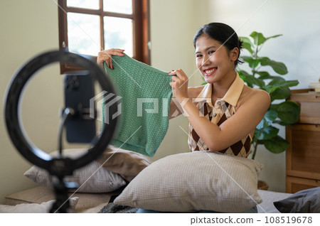 A beautiful female influencer or online seller is showing and reviewing her clothes to the camera. A beautiful female influencer or online seller is showing and reviewing her clothes to the camera. 108519678