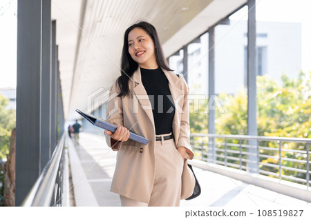An attractive Asian businesswoman is holding a binder while walking on a skywalk in the city. An attractive Asian businesswoman is holding a binder while walking on a skywalk in the city. 108519827
