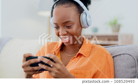 Phone games, headphones and woman in home on sofa for playing online gaming, podcast subscription or connection. Happy african person, video game or smartphone in living room for streaming multimedia Phone games, headphones and woman in home on sofa for playing online gaming, podcast subscription or connection. Happy african person, video game or smartphone in living room for streaming multimedia 108520493