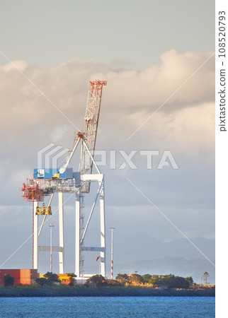 A crane and shipping containers in a shipyard surrounded by the ocean. Industrial hoisting crane in a logistics or cargo harbor at a seaport for the transportation of liquid materials A crane and shipping containers in a shipyard surrounded by the ocean. Industrial hoisting crane in a logistics or cargo harbor at a seaport for the transportation of liquid materials 108520793