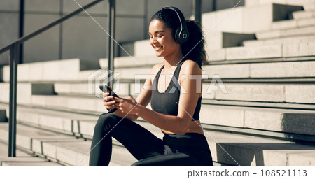 Runner girl, headphones and phone on stairs with music, smile and relax in city, workout and training. Woman, smartphone and sitting with audio streaming subscription with typing, social media or app Runner girl, headphones and phone on stairs with music, smile and relax in city, workout and training. Woman, smartphone and sitting with audio streaming subscription with typing, social media or app 108521113