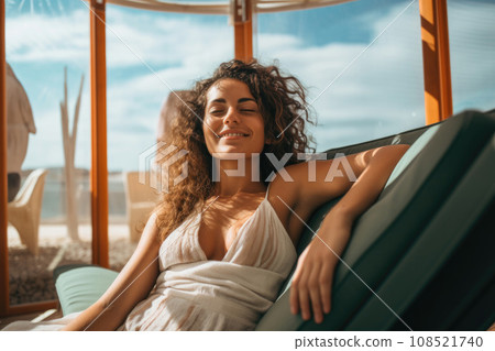 A young curly-haired woman lies with her eyes closed in a chaise longue on the seashore and smiles 108521740