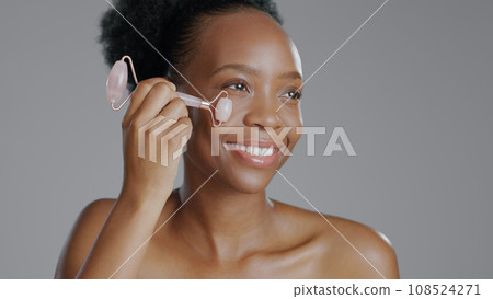 Facial roller, woman and smile in face massage in skincare or beauty, health and wellness or dermatology. Happy black person, skin and cosmetics with glow, care and tool in studio by gray background 108524271