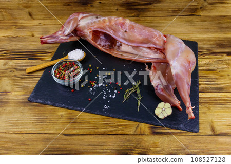 Whole raw rabbit meat with spices on a black slate cutting board on wooden table 108527128