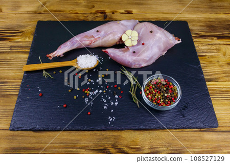 Raw rabbit legs with spices on a black slate cutting board on wooden table 108527129