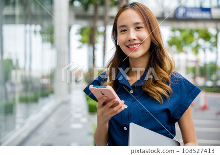 Young woman in blue shirt smiles while checking her smartphone on a city street. She is a professional, using her device to stay connected to her work and business. 108527413
