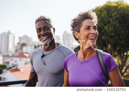 Adult international couple in sports outfits looking at camera with energetic cheerful smile. Happy loving mature man and woman jogging or exercising outdoors. Healthy lifestyle in urban environment. Adult international couple in sports outfits looking at camera with energetic cheerful smile. Happy loving mature man and woman jogging or exercising outdoors. Healthy lifestyle in urban environment. 108528702