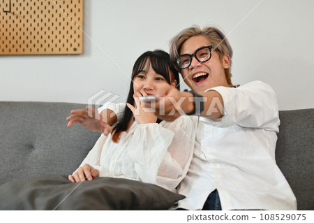 Joyful young couple with remote control sitting on sofa at home watching television on weekend Joyful young couple with remote control sitting on sofa at home watching television on weekend 108529075