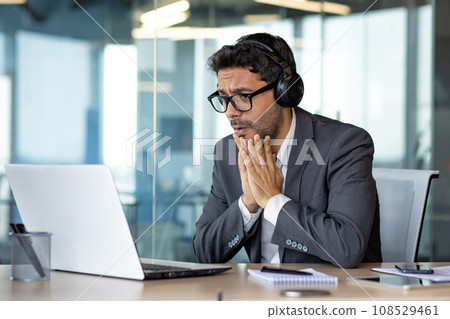Man watching sports match inside office at workplace, cheering, businessman in headphones and laptop sitting at table, unhappy with result sad and disappointed. 108529461