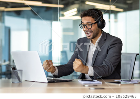 A man at the workplace inside the office is watching a sports match, a businessman in headphones and with a laptop is cheering at the workplace, a joyful boss, celebrating a victory, smiling. 108529463