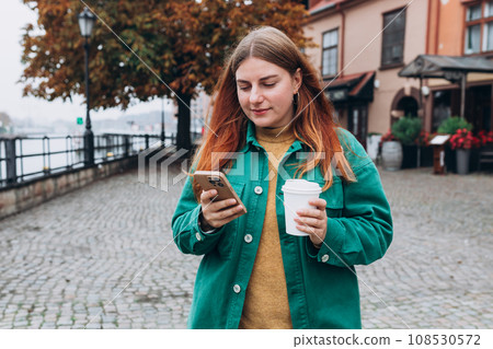 Happy cheerful young woman with paper coffee cup walking on city street checks her smartphone. Portrait of beautiful 30s girl using smartphone outdoors. Urban lifestyle concept. Happy cheerful young woman with paper coffee cup walking on city street checks her smartphone. Portrait of beautiful 30s girl using smartphone outdoors. Urban lifestyle concept. 108530572