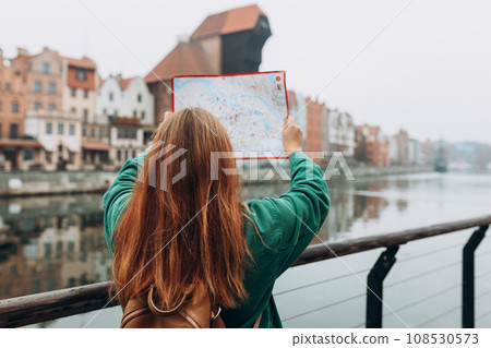 Attractive young female tourist is exploring new city. Redhead girl with backpack holding a paper map on city street in Gdansk. Traveling Europe in autumn. Famous Zuraw crane, Motlawa river Attractive young female tourist is exploring new city. Redhead girl with backpack holding a paper map on city street in Gdansk. Traveling Europe in autumn. Famous Zuraw crane, Motlawa river 108530573