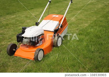 Top view of modern orange-grey electric lawn mower cutting bright lush green grass. Gardening work tools. Rotary lawn mower machine on lawn. Professional lawn care service. Place for text 108530903