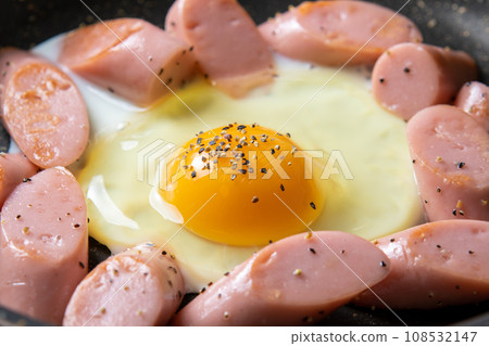 A cooking scene where fried eggs (with black pepper) and fish sausage are fried in a frying pan. A cooking scene where fried eggs (with black pepper) and fish sausage are fried in a frying pan. 108532147