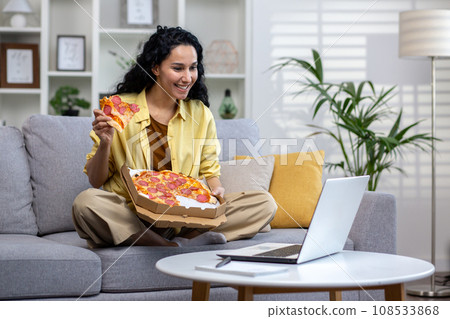 Joyful and happy woman eating pizza alone at home, Hispanic woman satisfied with timely food delivery, using laptop to watch online video stream, sitting on sofa in living room at home. 108533868