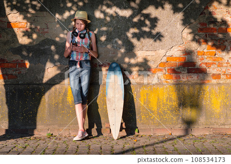 Portrait of a young woman with a skateboard leaning against a wall in the city 108534173
