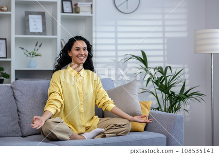 Young beautiful woman meditating at home sitting on sofa in living room in lotus pose, latin american joyful smiling, dreaming and visualizing future achievement results and plans. 108535141