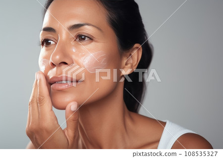 Close-up of middle-aged Caucasian woman touching her face to apply moisturizer. Smiling face of adult brunette lady with daily cream, facial cosmetics. Skin care. Grey background, copy space. Close-up of middle-aged Caucasian woman touching her face to apply moisturizer. Smiling face of adult brunette lady with daily cream, facial cosmetics. Skin care. Grey background, copy space. 108535327