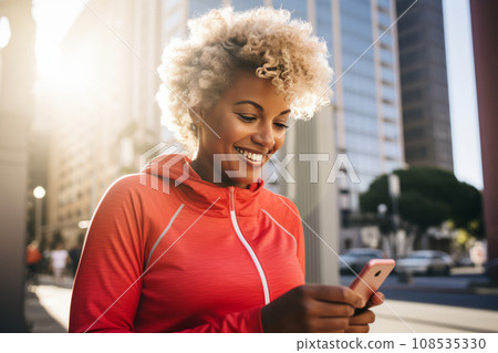 Young athletic African American woman in sportswear with smartphone while training outdoor. Slender black lady resting after jogging or workout in city street. Active lifestyle in urban environment. Young athletic African American woman in sportswear with smartphone while training outdoor. Slender black lady resting after jogging or workout in city street. Active lifestyle in urban environment. 108535330