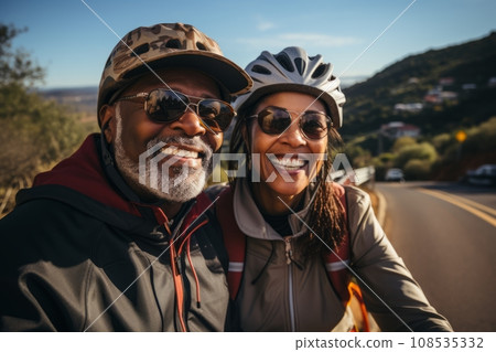 Portrait of elderly smiling African American couple riding bicycles together along picturesque country road. Cheerful seniors on a bike ride. Retired people lead active lifestyle to stay healthy. Portrait of elderly smiling African American couple riding bicycles together along picturesque country road. Cheerful seniors on a bike ride. Retired people lead active lifestyle to stay healthy. 108535332