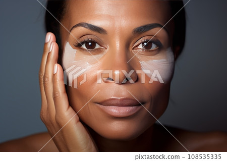 Close-up of middle-aged African American woman touching her face to apply moisturizer. Smiling face of adult colored lady with daily cream, facial cosmetics. Skin care. Grey background, copy space. Close-up of middle-aged African American woman touching her face to apply moisturizer. Smiling face of adult colored lady with daily cream, facial cosmetics. Skin care. Grey background, copy space. 108535335