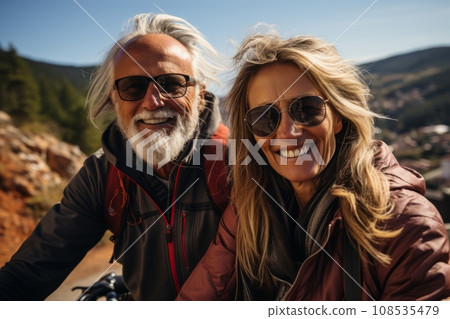Close-up portrait of elderly smiling Caucasian couple riding bicycles together along picturesque country road. Cheerful seniors on a bike ride. Retired people lead active lifestyle to stay healthy. Close-up portrait of elderly smiling Caucasian couple riding bicycles together along picturesque country road. Cheerful seniors on a bike ride. Retired people lead active lifestyle to stay healthy. 108535479