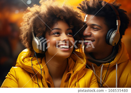 Close up of cheerful African American couple in yellow jackets enjoying favorite playlist in headphones. Happy smiling young people listening to music or podcast in app, having fun and laughing. Close up of cheerful African American couple in yellow jackets enjoying favorite playlist in headphones. Happy smiling young people listening to music or podcast in app, having fun and laughing. 108535480