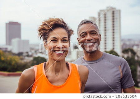Adult international couple in sports outfits looking at camera with energetic cheerful smile. Happy loving mature man and woman jogging or exercising outdoors. Healthy lifestyle in urban environment. Adult international couple in sports outfits looking at camera with energetic cheerful smile. Happy loving mature man and woman jogging or exercising outdoors. Healthy lifestyle in urban environment. 108535504
