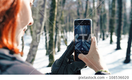 Young woman with gloves taking snow pictures with her phone  108536020