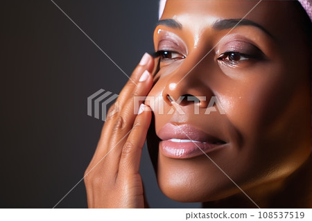Close-up of middle-aged African American woman touching her face to apply moisturizer. Smiling face of adult colored lady with daily cream, facial cosmetics. Skin care. Grey background, copy space. 108537519