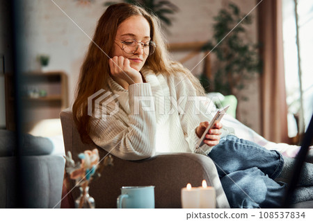 Attractive young girl sitting on sofa at home and talking on phone video call. Comfort and cozy home atmosphere. Attractive young girl sitting on sofa at home and talking on phone video call. Comfort and cozy home atmosphere. 108537834