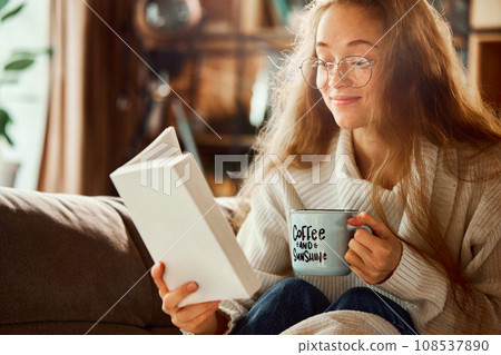 Portrait of young beautiful woman, student in glasses reading favorite funny story, book sitting on sofa at home. 108537890